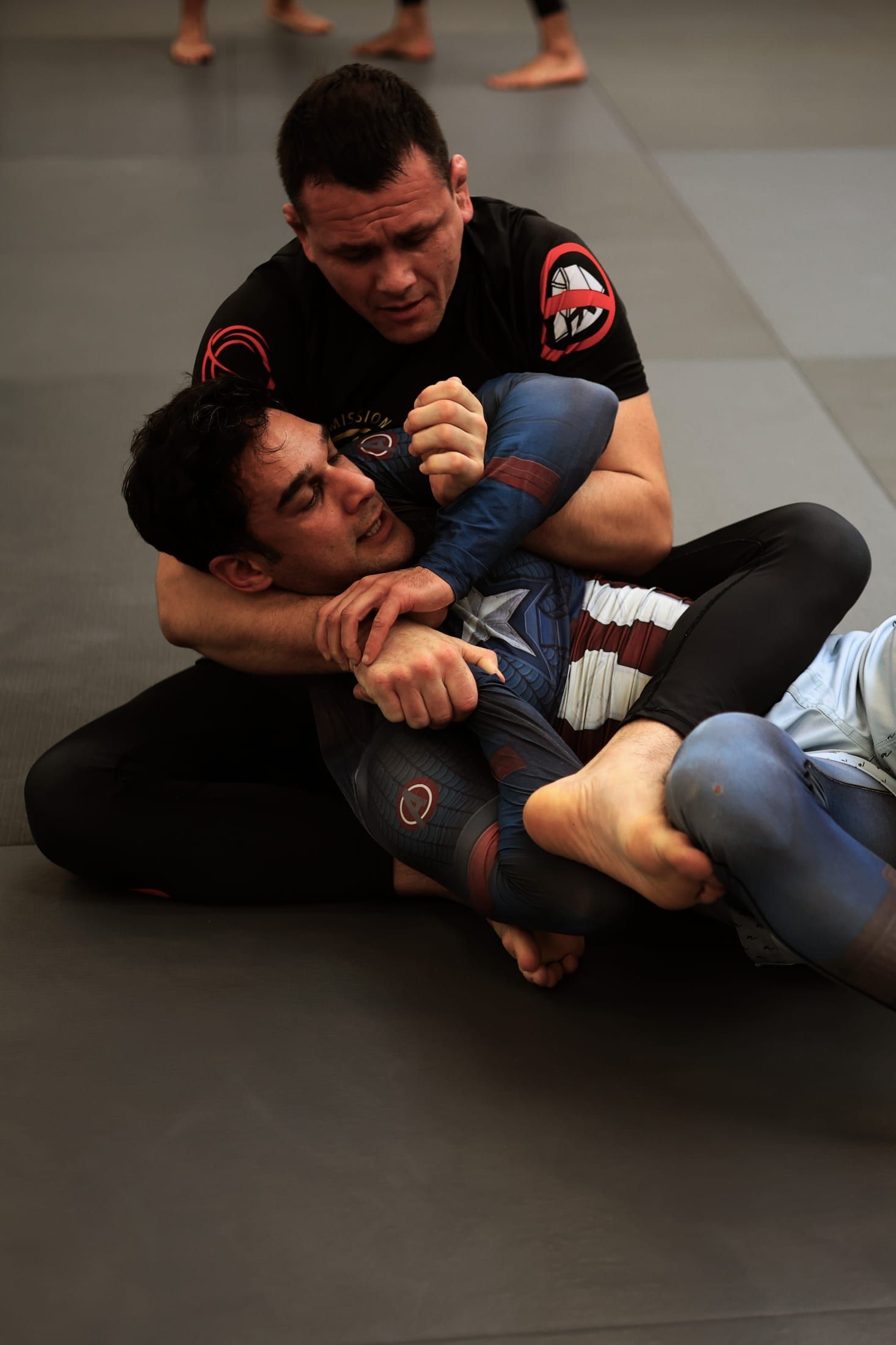 Gurinder Sandhu rolling no-gi on the mats, hands framing against a training partner’s arm across his neck